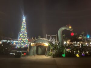 Large dinosaur statue holding a lit Christmas ornament in its mouth and wearing a Santa hat, in front of a tall full lit Christmas tree at Walt Disney World's Hollywood Studios park